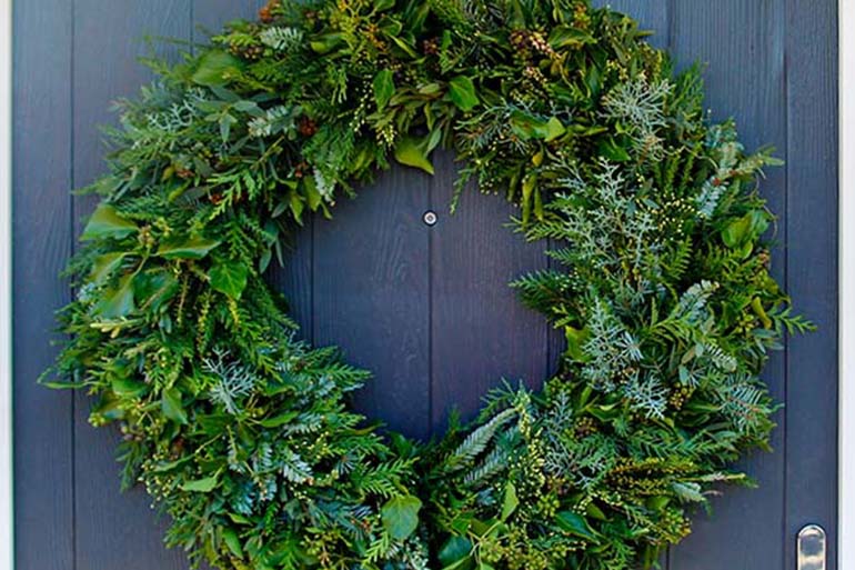 evergreen foliage on a table, with a pair of hands wearning blue gloves picking through it to attach it to a metal wreath ring.